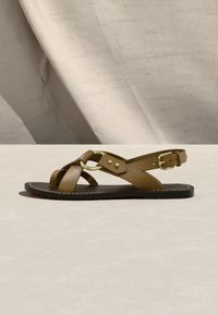 Brūns ādas plakanas sandāls ar krustotām siksniņām, zelta sprādzi un gredzena detaļām, izvietots uz bēša virsmas ar auduma fonu.