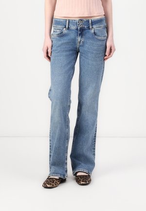 Personne portant un jean droit bleu clair, un haut court côtelé rose, et des chaussures plates à imprimé léopard, debout sur un sol blanc.