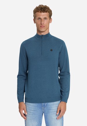 Mann, der einen blauen Viertel-Reißverschluss-Pullover mit kleinem schwarzem Emblem auf der Brust und hellblaue Jeans trägt, steht vor weißem Hintergrund.