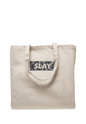 Béžová plátená taška s dvoma rukoväťami a čiernou obdĺžnikovou nášivkou so vzormi paisley v bielej farbe a slovom „SLAY“ v tučnom písme.