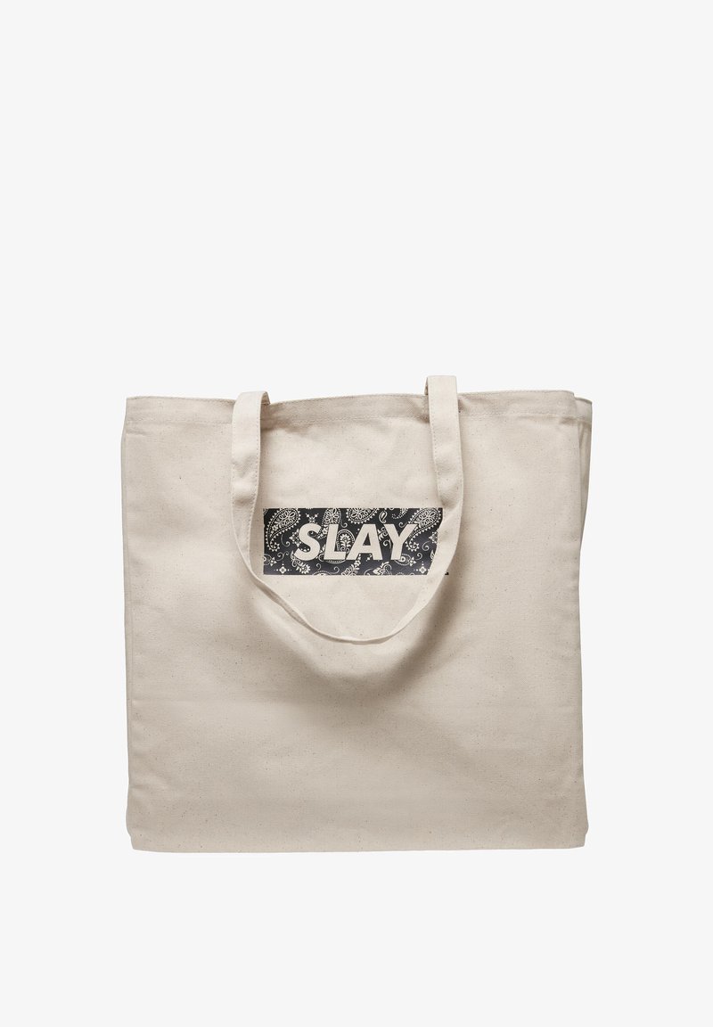 Sac cabas en toile beige avec deux anses et un patch rectangulaire noir présentant des motifs paisley blancs et le mot « SLAY » en lettres grasses.