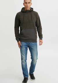 Sudadera de forro polar verde oscuro con media cremallera, bolsillo frontal gris y cordones naranjas, combinada con jeans azul claro y zapatillas negras.