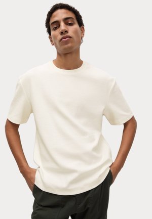 White short-sleeve shirt in textured fabric, crew neckline, relaxed fit. Paired with dark green pants, hands in pockets.