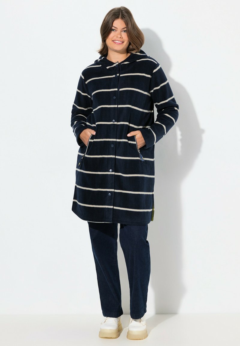 Cappotto con cappuccio a righe navy con linee orizzontali di un beige chiaro, chiusura con bottoni, due tasche laterali, realizzato in tessuto morbido, abbinato a jeans scuri e scarpe.