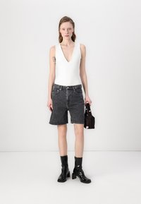 Bodysuit blanc à col en V profond associé à un short en denim noir taille haute. Le modèle porte des bottes noires vernies et tient un petit sac noir.