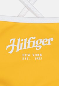 Yellow swimsuit with white trim featuring "Hilfiger" logo and "NEW YORK EST. 1985" text in bold typography. Smooth, textured fabric.