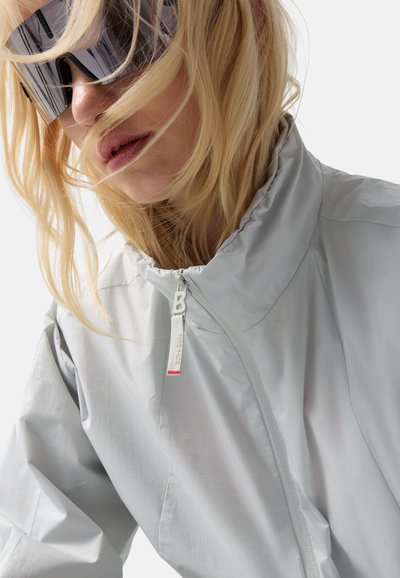 Blond persoon met los haar, draagt grote zwarte zonnebril en een lichtgrijs jack met rits en hoge kraag.