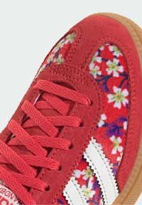 Roter Wildleder-Sneaker mit einer seitlichen Blütenstoffpaneele, weißen Lederakzenten, strukturierten Schnürsenkeln und einer Gummisohle für besseren Halt.