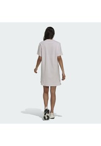 Jasnoróżowa, oversize sukienka t-shirt z krótkimi rękawami i okrągłym dekoltem, wykonana z gładkiej bawełny, sięgająca do połowy uda, noszona z białymi trampkami.