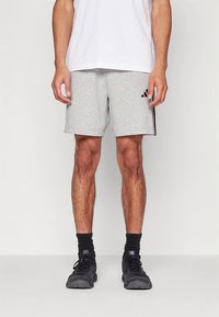 Calções desportivos cinzentos feitos de tecido macio, com riscas pretas nas laterais e um logótipo Adidas na perna esquerda. Estilizados com uma t-shirt branca.