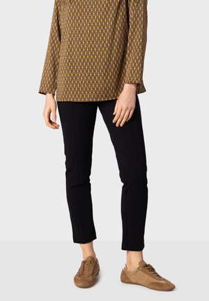Personne portant un pantalon noir slim, un haut marron à manches longues à motif, et des chaussures en daim marron à lacets, debout devant un fond uni.
