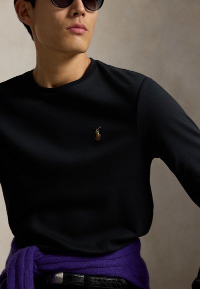 Mand iført sort langærmet skjorte med lille broderet polo-spiller logo, lilla sweater bundet om livet og mørke solbriller.