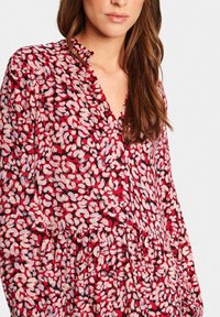 Punaiset ja vaaleanpunaiset kukkakuosiset puserot, joissa on V-pääntie, röyhelödetaljit, pitkät hihat ja kerätty vyötärö, valmistettu kevyestä kankaasta.