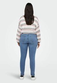 Šviesiai mėlynos skinny džinsai su aukštu liemeniu, derinami su balta, daugiaspalve dryžuota megztiniu. Megztinis turi ilgomis rankovėmis ir laisvą siluetą.