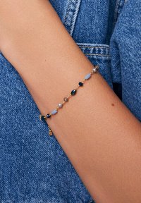 Złota bransoletka z różnorodnymi niebieskimi, zielonymi i neutralnymi koralikami w różnych kształtach i rozmiarach, z małym zawieszką.