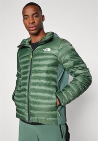 Veste rembourrée verte avec capuche, dotée d'une fermeture éclair à l'avant, de poches latérales et d'un logo sur la poitrine. Tissu texturé et isolé.