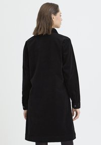 Rochie cămașă din catifea neagră, cu mâneci lungi, mansete cu nasturi și o siluetă dreaptă. Prezintă un guler și o lungime până la genunchi. Textură netedă.