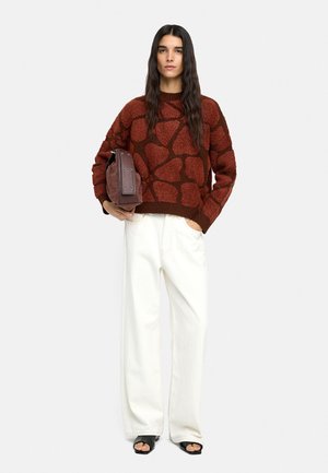 Kobieta z długimi ciemnymi włosami, ubrana w czerwony sweter w wzory, białe spodnie o szerokich nogawkach, czarne buty na obcasie z odkrytymi palcami, trzyma złożoną brązową torbę, stoi.