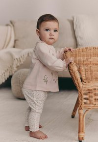 Top de manga larga en rosa claro con bordado floral y puños con volantes, combinado con leggings blancos con un pequeño estampado floral. Silla natural de mimbre.