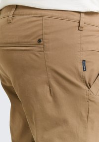 Tan broek met een gladde textuur, voorzien van een knopen achterzak en een klein label aan de zijkant, met een klassieke uitstraling.