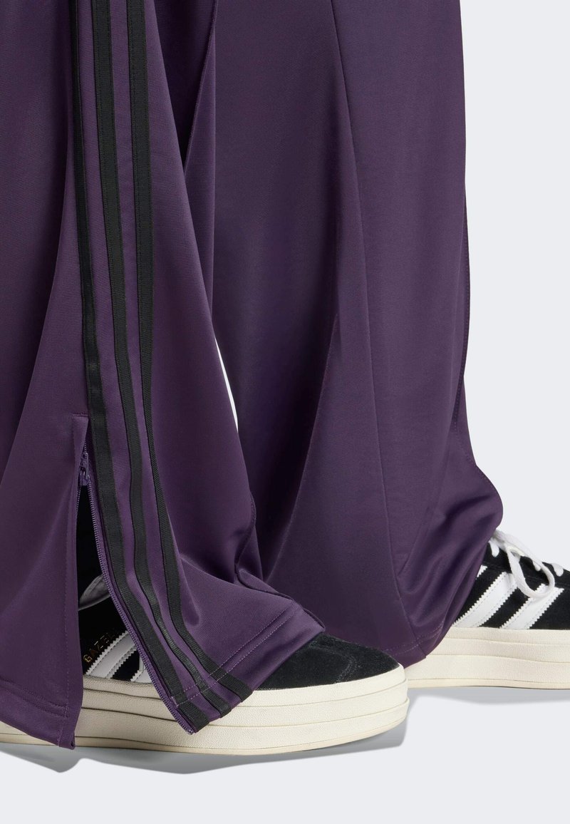 Pantalons de sport violets avec des bandes noires sur les côtés et des zips à l'ourlet, associés à des baskets noir et blanc avec des semelles compensées.