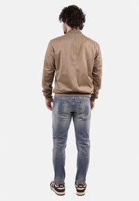 Giacca bomber marrone chiaro con una texture liscia, abbinata a jeans blu sbiaditi con dettagli sottili e sneaker marroni con accenti bianchi.