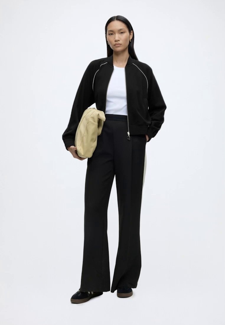 Femme portant une veste noire zippée, chemise blanche, pantalon large noir, tenant un sac beige, debout devant un fond clair uni.