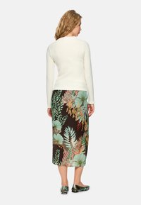 Langärmliges cremefarbenes Oberteil mit Struktur, kombiniert mit einem schwarzen Rock mit floralen Mustern in Grün und Orange; glänzende dunkelgrüne Ballerinas.