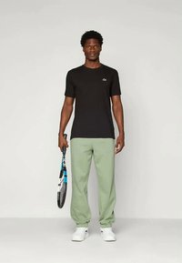 Vīrietis, kas valkā melnu T-kreklu, gaiši zaļas sporta bikses un baltas sporta apavus, tur tenisista raketi un stāv pret vienkāršu gaišu fona.