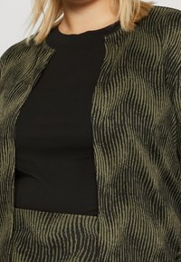 Vzorovaná oliva zelená bunda s černými pruhy, s vysokým límcem a otevřeným předním dílem, vrstvená přes jednobarevný černý top.