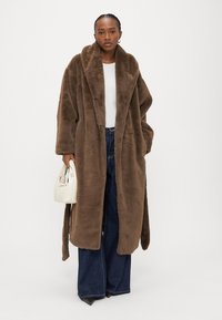 Žena v dlhom hnedom umelom kožuchu, bielom topu, širokých tmavých džínsach, drží malú bielu kabelku, stojí na bielom pozadí.