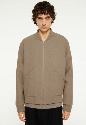 Jeune homme aux cheveux bouclés portant une veste bomber beige zippée par-dessus un pull et un pantalon noir sur un fond clair uni.