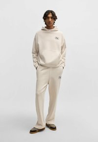 Felpa beige con cappuccio e pantaloni coordinati realizzati in tessuto morbido, con una tasca frontale e dettagli del logo neri. Calzature marroni e nere completano il look.