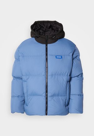 Lichtblauwe pufferjack met zwarte capuchon, ritssluiting aan de voorkant en horizontale stiksels. Voorzien van een logo in het blauw op de linkerborst.