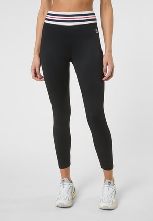 Mujer con mallas negras acanaladas con una cinturilla a rayas roja, blanca y azul, y zapatillas blancas con detalles en gris.