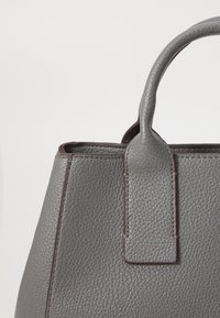 Sac à main en cuir gris avec une finition texturée, une anse robuste et une bordure foncée. Présente un accent rectangulaire près de la anse.