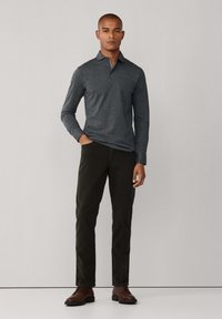 Camisa polo de manga larga gris oscuro, con cuello abotonado, combinada con pantalones verde oscuro y zapatos marrones de cordones sobre un fondo neutro.