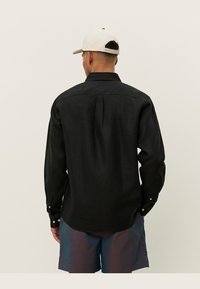 Zwarte langemouwen shirt van textuurstof, met een button-down kraag, rugplooien en knoopmanchetten. Draag het met kleurrijke shorts.