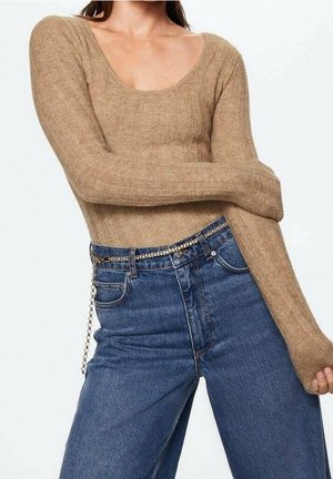 Femme portant un haut à manches longues côtelé beige et un jean taille haute bleu avec une fine ceinture en chaîne, posant les bras croisés.