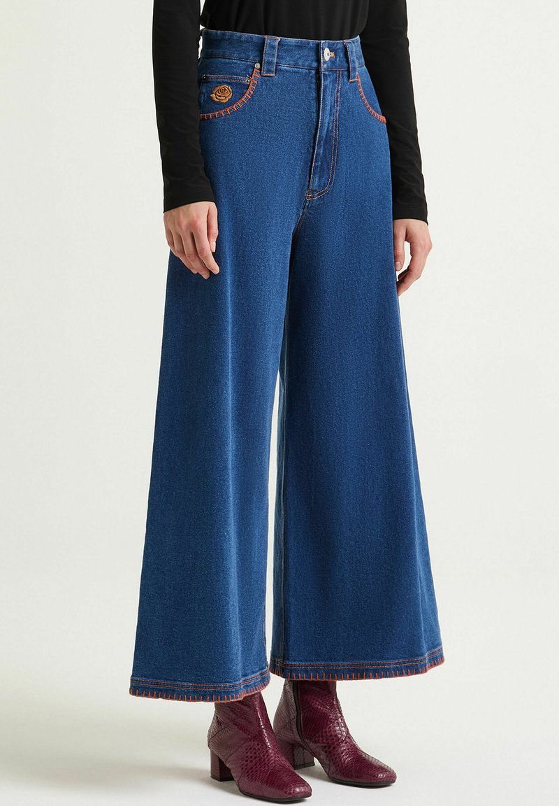 Pantaloni di jeans a gamba larga in blu scuro con cuciture arancioni, caratterizzati da tasche decorative anteriori e vita alta, abbinati a stivaletti bordeaux.