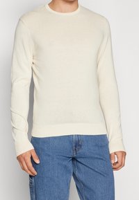 Homme portant un pull à manches longues de couleur crème associé à un jean en denim bleu, debout devant un fond clair uni.