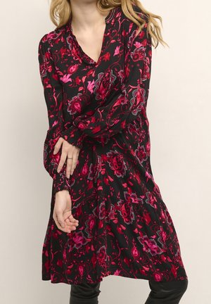 Frau in einem schwarzen Kleid mit langen Ärmeln und einem lebhaften roten und pinken Blumen-Paisley-Muster, die mit vor der Brust verschränkten Armen posiert.