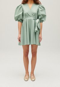 Zelené, krátké zavinovací šaty s nabíranými rukávy, texturovaná látka, detaily se zavázaným pasem a rozšířená sukně. Doplněné o zlaté ploché boty.