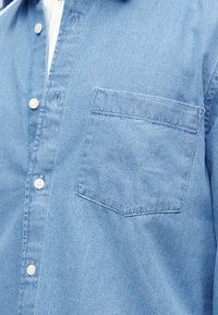 Ανοιχτό μπλε τζιν πουκάμισο με λευκά κουμπιά και μία τσέπη στο στήθος, φορεμένο πάνω από λευκό φανελάκι.