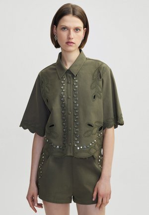 Chemise à manches courtes vert olive avec des motifs floraux brodés et des boutons décoratifs sur le devant, présentant des bords festonnés.