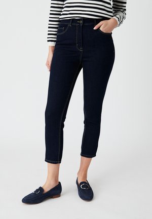 Jeans de mezclilla azul oscuro con un diseño ajustado, largo recortado, costuras doradas y bolsillos frontales, combinados con mocasines de ante azul marino.