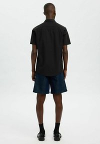 Camisa negra de manga corta con dobladillo recto, combinada con pantalones cortos de mezclilla oscura hasta la rodilla. El modelo lleva calcetines y zapatos negros.