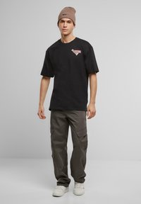 Black cotton t-shirt with short sleeves and a colorful graphic pattern on the chest, paired with grey cargo pants and a beige beanie.