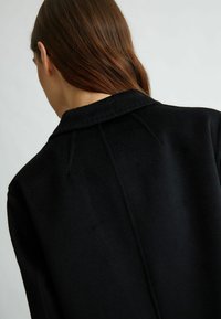 Cappotto di lana nera con colletto a punte, texture liscia e dettagli delle cuciture sottili, che mette in mostra un design minimalista.