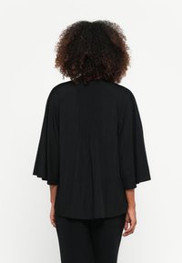 Blusa preta com mangas largas em forma de sino, tecido texturizado e corte solto. Apresenta um design com costura franzida nas costas e um decote arredondado.